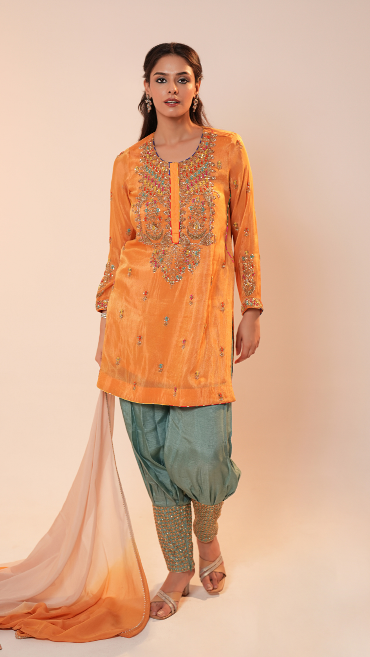 Woman wearing an orange embroidered kurta with green pants on a beige background