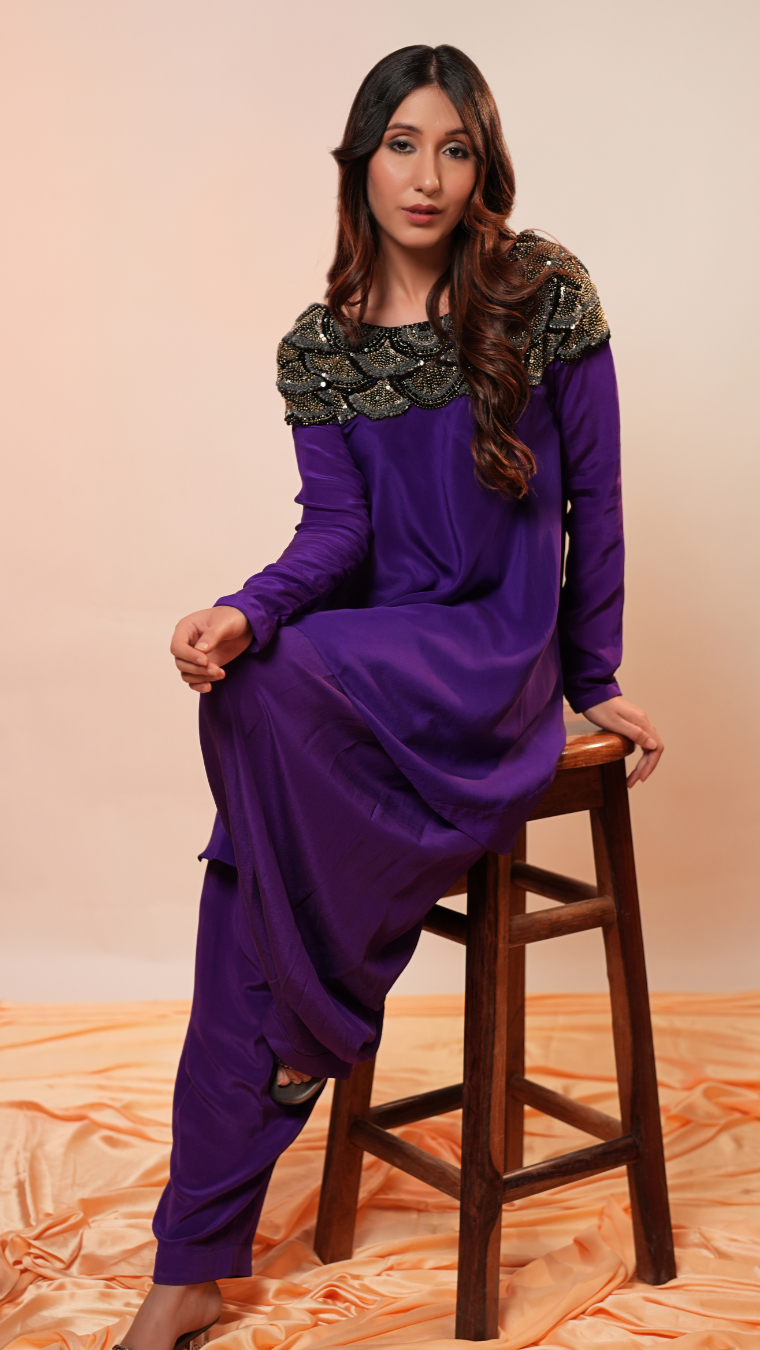 Woman in a purple outfit with green embellishments sitting on a wooden stool against an orange fabric background.