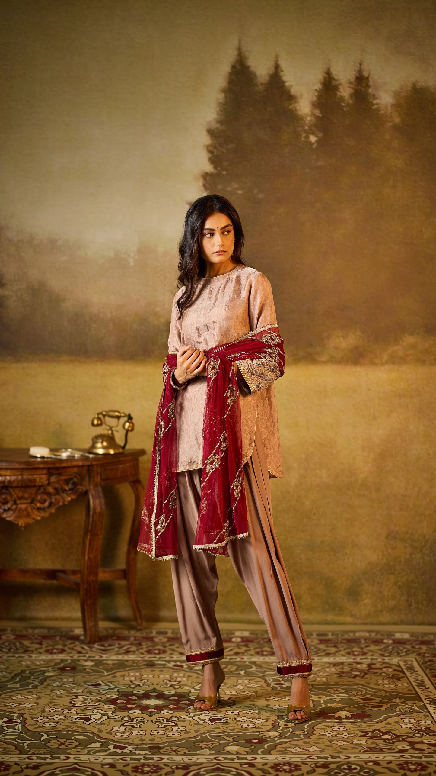 Woman in traditional outfit standing in a room with a painting of a landscape in the background