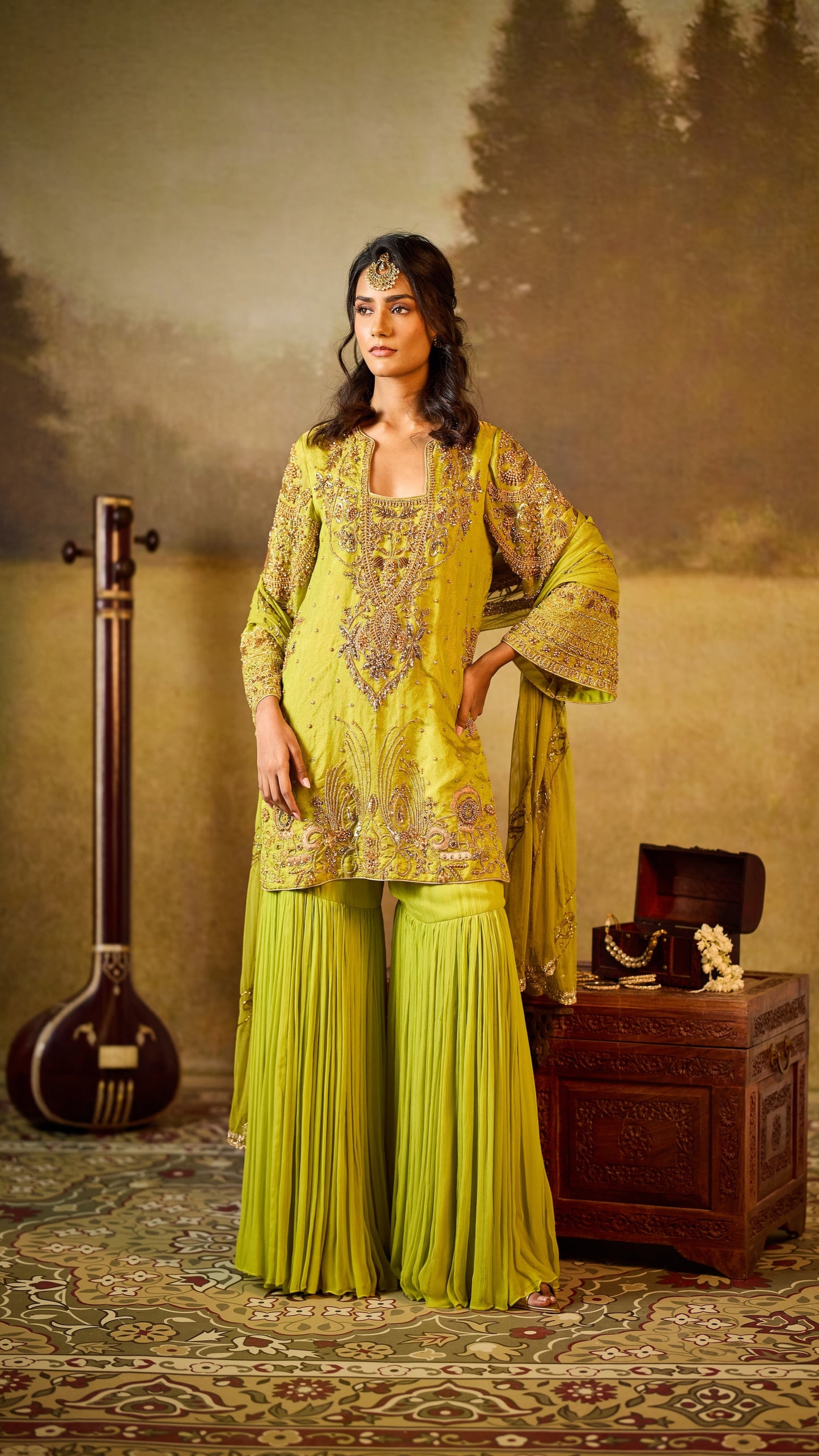 Woman in a green traditional outfit standing in a decorated room with a musical instrument and wooden chest.