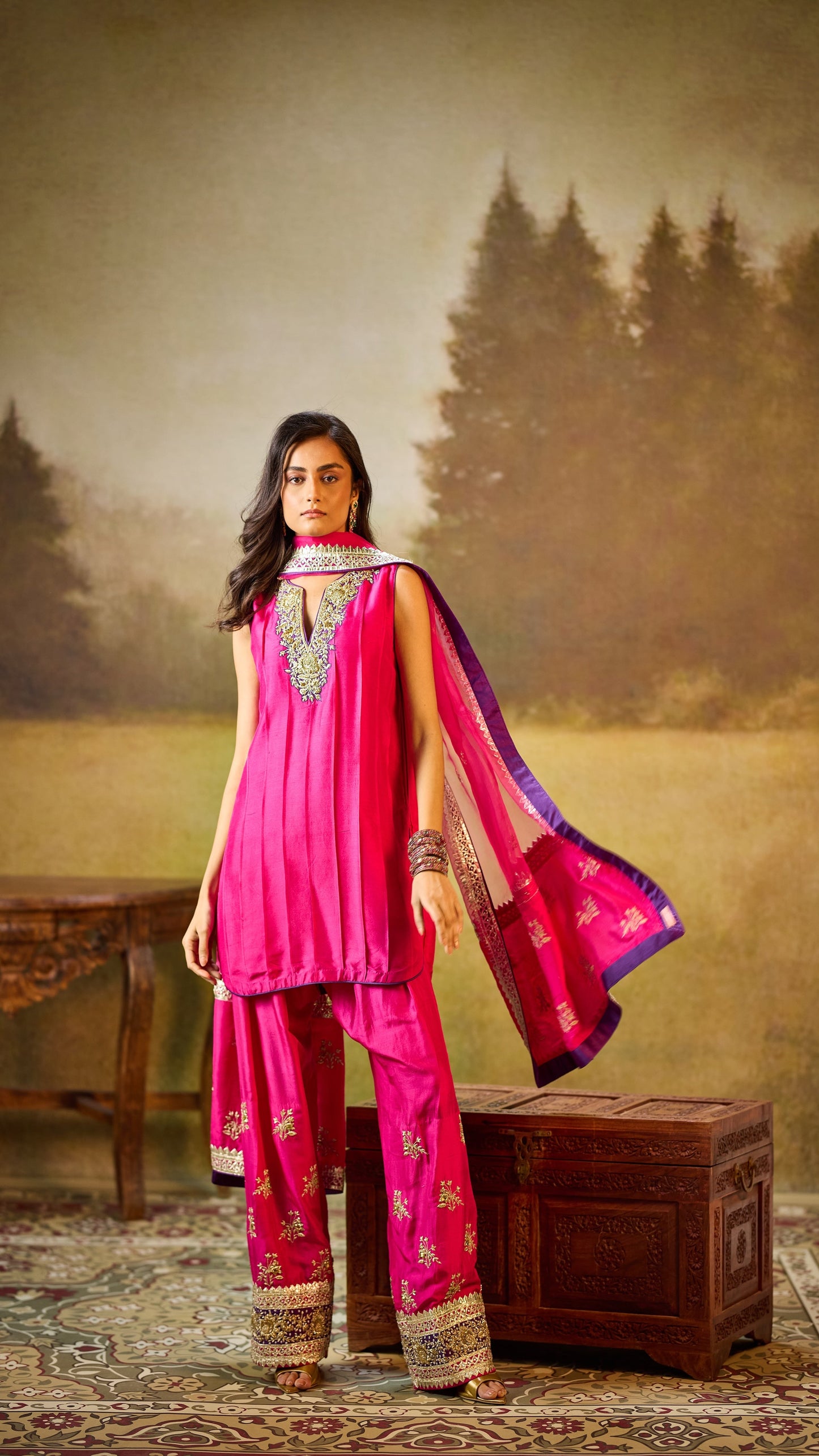 Woman in a pink traditional outfit standing next to a wooden trunk with a misty forest background.
