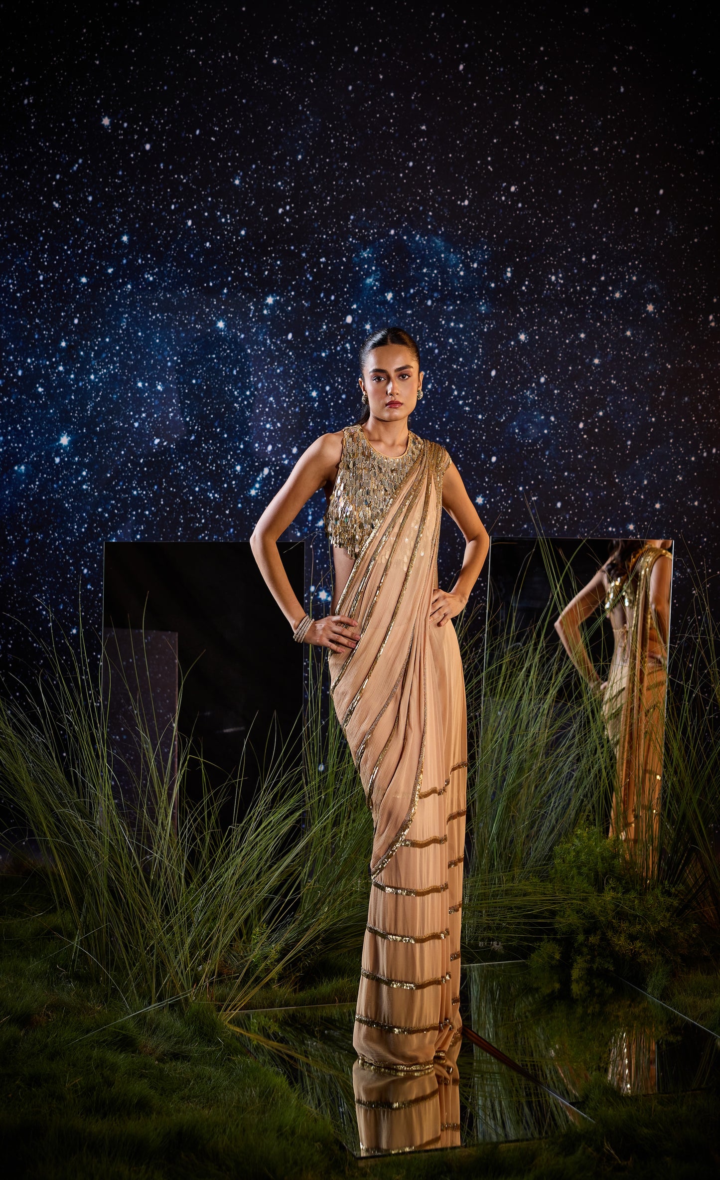 Woman in a saree standing in front of a starry night sky with grass and water below.