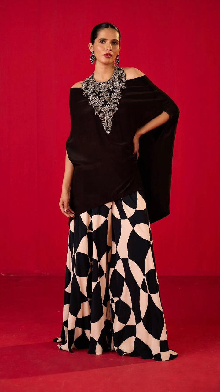 A model wearing an off-shoulder black top with a geometric patterned sharara, standing against a red background.