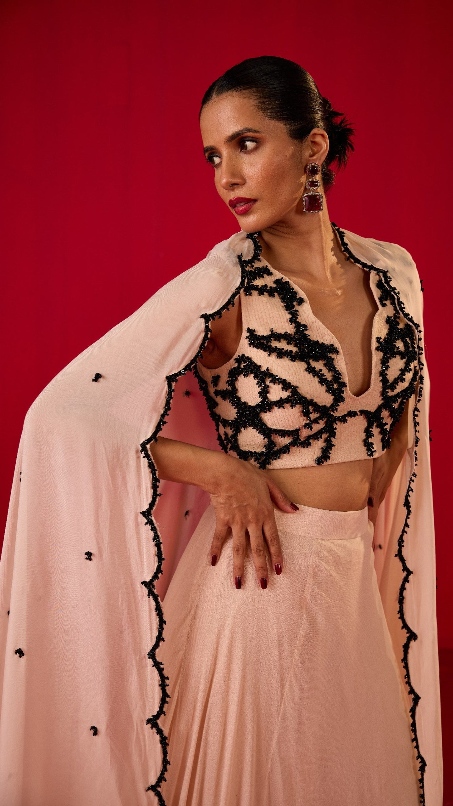 A woman posing in a traditional pink lehenga with black embellishments and a dupatta, against a red background.