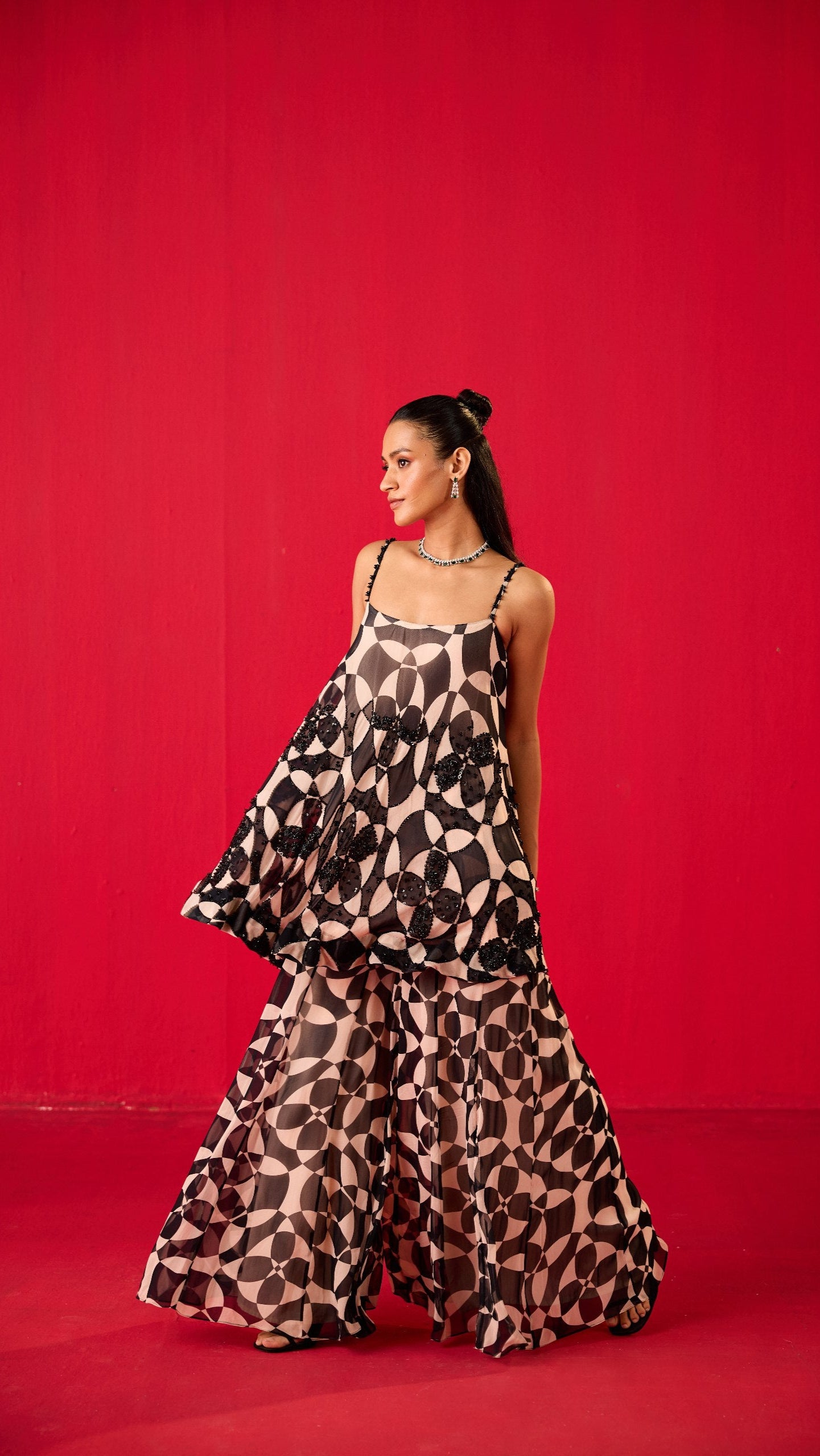 A woman standing against a red background, wearing a traditional Sharara with a leopard print pattern and a floral belt.