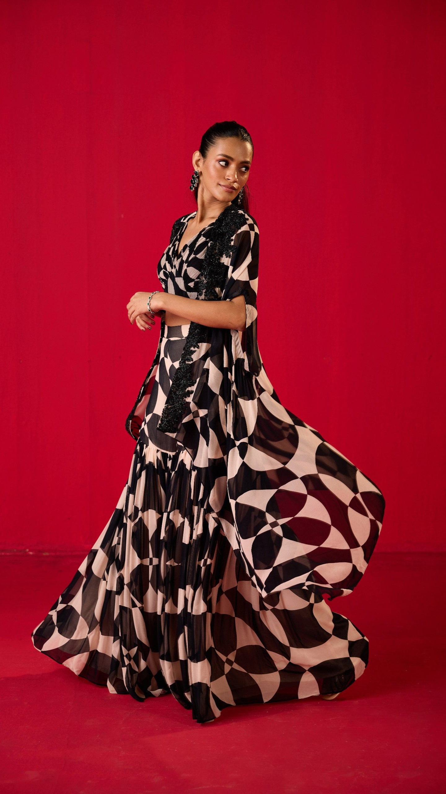 A woman wearing a traditional, flowing sharara with a geometric pattern, predominantly in black and white colors, posing against a red background.