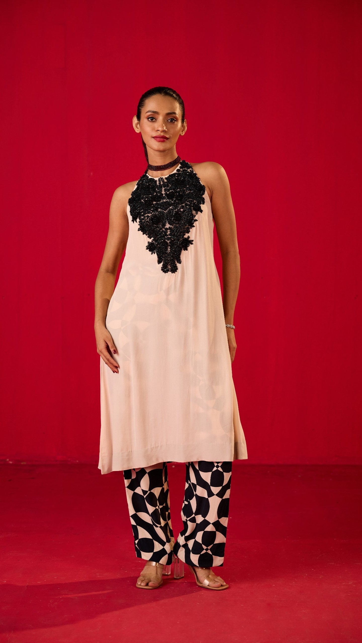 A woman standing against a red background, wearing a traditional Kurta set with a black top and white skirt, accessorized with a black belt and necklace.