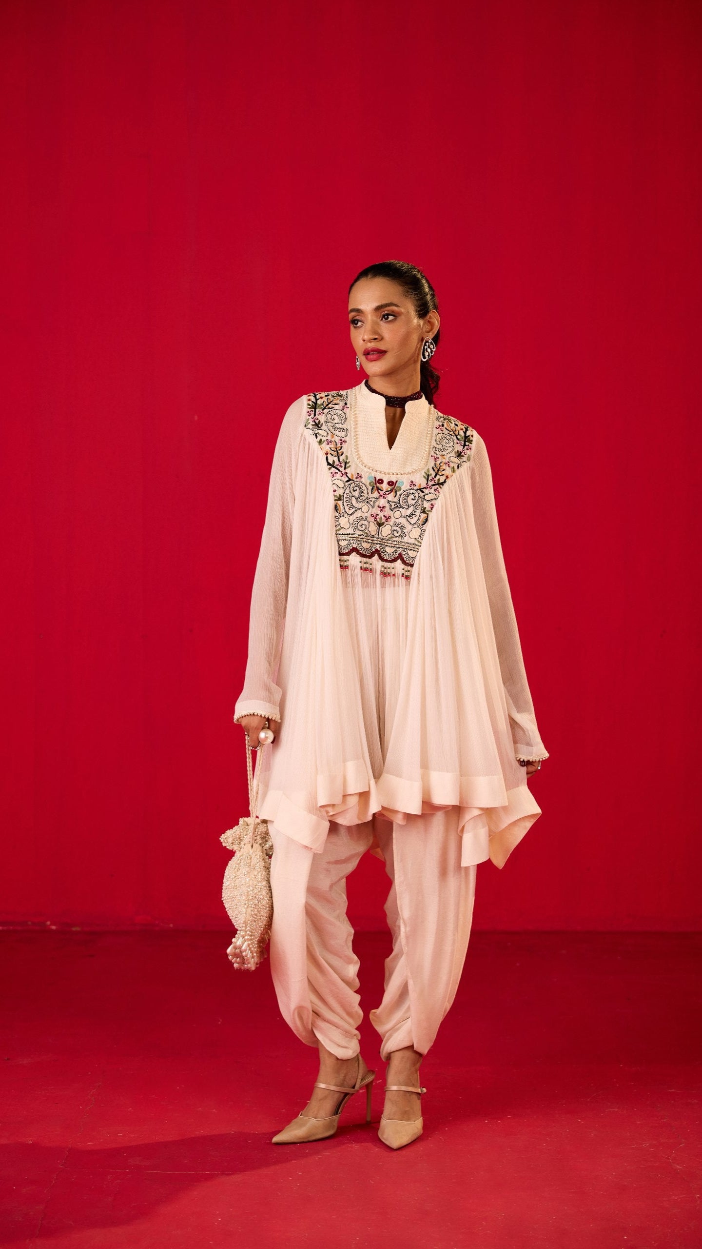 A model wearing an off white long top with lungi salwar, featuring embroidery on the chest area, standing against a red background.
