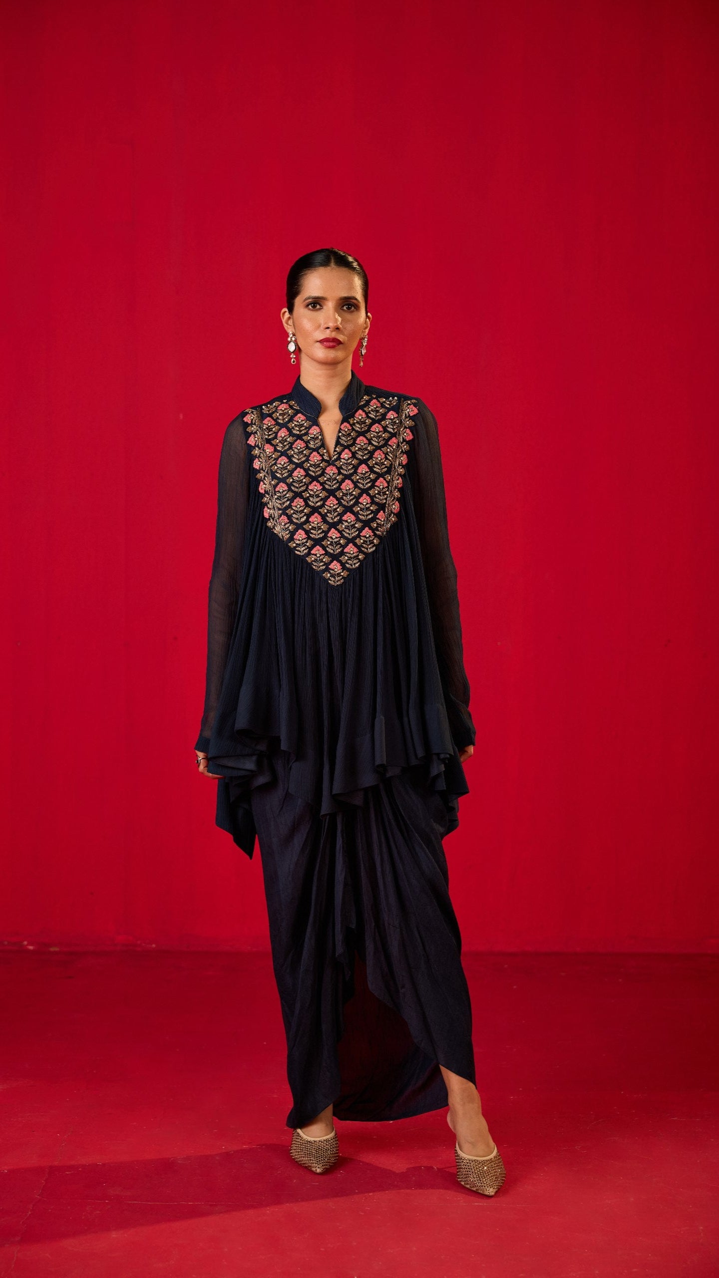 A person wearing a navy blue loose kurta with lungi skirt standing against a red background.